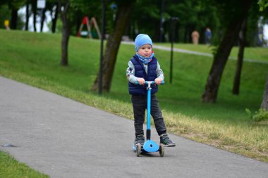 Yeşil bir parkta mavi bir scooter 'a binen, sıcak giysili küçük bir çocuk. Çocuk ilkbahar ya da sonbaharın başında açık havada eğleniyor. Düzgün bir kaldırımda güvenli ve eğlenceli bir gezinti..