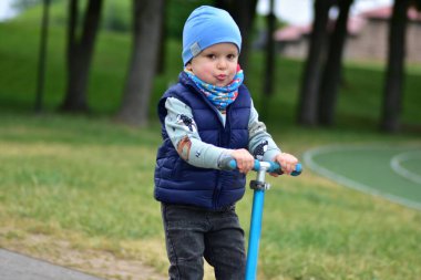 Yeşil bir parkta mavi bir scooter 'a binen, sıcak giysili küçük bir çocuk. Çocuk ilkbahar ya da sonbaharın başında açık havada eğleniyor. Düzgün bir kaldırımda güvenli ve eğlenceli bir gezinti..