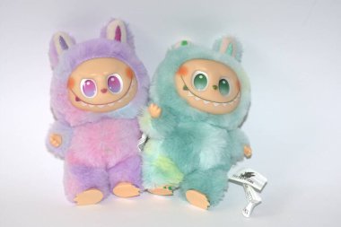 Kabarık yeşil ve pembe canavar kostümlü iki oyuncu Labubu oyuncak figürü ve beyaz bir arka plana karşı muzip bir gülümseme..