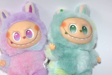Kabarık yeşil ve pembe canavar kostümlü iki oyuncu Labubu oyuncak figürü ve beyaz bir arka plana karşı muzip bir gülümseme..