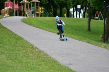 Yeşil bir parkta mavi bir scooter 'a binen, sıcak giysili küçük bir çocuk. Çocuk ilkbahar ya da sonbaharın başında açık havada eğleniyor. Düzgün bir kaldırımda güvenli ve eğlenceli bir gezinti..