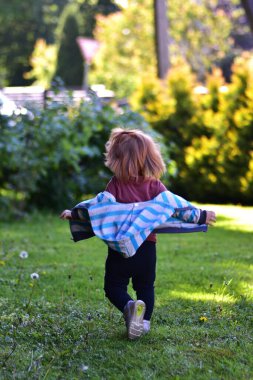 Güneşli bir günde kolları açık, elinde çizgili bir ceketle dışarı koşan mutlu kızıl saçlı küçük kız. Kaygısız çocukluk anı canlı yeşil bir bahçede sıcacık doğal ışıkla yakalanır..