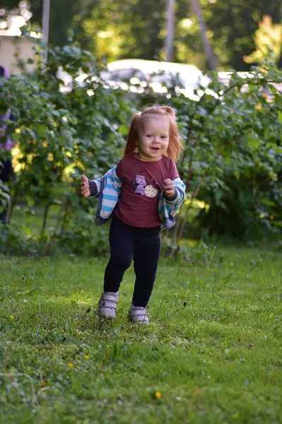 Güneşli bir günde elinde çizgili bir ceketle dışarı koşan mutlu kızıl saçlı kız. Kaygısız çocukluk anı canlı yeşil bir bahçede sıcacık doğal ışıkla yakalanır..