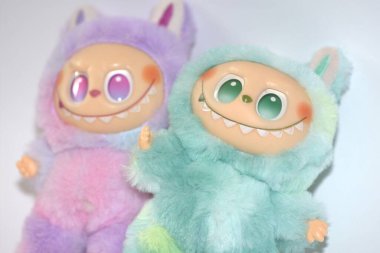 Kabarık yeşil ve pembe canavar kostümlü iki oyuncu Labubu oyuncak figürü ve beyaz bir arka plana karşı muzip bir gülümseme..