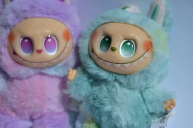 Kabarık mavi ve pembe canavar kostümlü iki oyuncu Labubu oyuncak figürü ve doğrudan güneş ışığı altında beyaz bir arkaplan karşısında muzip bir gülümseme..