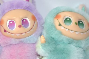 Kabarık yeşil ve pembe canavar kostümlü iki oyuncu Labubu oyuncak figürü ve beyaz bir arka plana karşı muzip bir gülümseme..