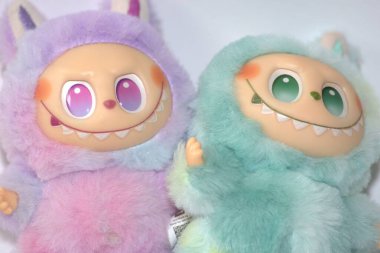 Kabarık yeşil ve pembe canavar kostümlü iki oyuncu Labubu oyuncak figürü ve beyaz bir arka plana karşı muzip bir gülümseme..