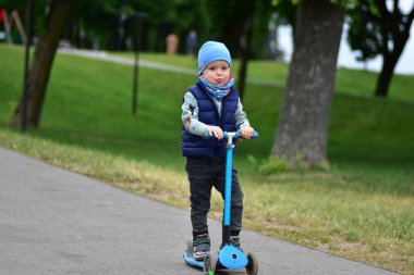 Yeşil bir parkta mavi bir scooter 'a binen, sıcak giysili küçük bir çocuk. Çocuk ilkbahar ya da sonbaharın başında açık havada eğleniyor. Düzgün bir kaldırımda güvenli ve eğlenceli bir gezinti..
