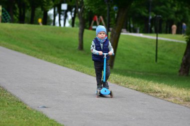 Yeşil bir parkta mavi bir scooter 'a binen, sıcak giysili küçük bir çocuk. Çocuk ilkbahar ya da sonbaharın başında açık havada eğleniyor. Düzgün bir kaldırımda güvenli ve eğlenceli bir gezinti..