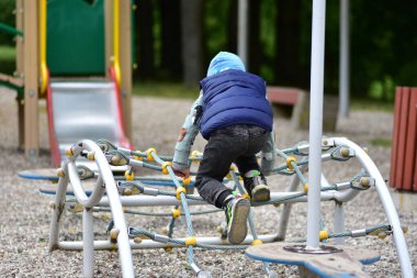 Küçük çocuk parktaki bir oyun parkında mavi yelek ve bere giymiş bir halat yapısına tırmanıyor. Güvenli bir açık alanda macera, oyun ve motor beceriler geliştirme ruhunu yakalar..