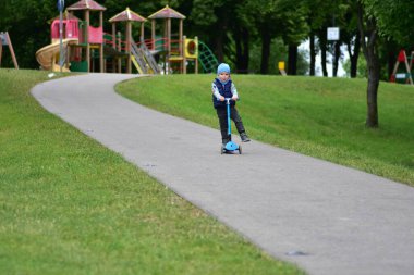 Yeşil bir parkta mavi bir scooter 'a binen, sıcak giysili küçük bir çocuk. Çocuk ilkbahar ya da sonbaharın başında açık havada eğleniyor. Düzgün bir kaldırımda güvenli ve eğlenceli bir gezinti..