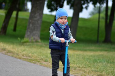 Yeşil bir parkta mavi bir scooter 'a binen, sıcak giysili küçük bir çocuk. Çocuk ilkbahar ya da sonbaharın başında açık havada eğleniyor. Düzgün bir kaldırımda güvenli ve eğlenceli bir gezinti..