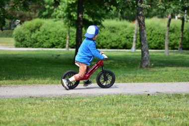 Park yolunda denge bisikleti süren küçük bir çocuk. Şapka takan ve spor kıyafetli çocuklar güneşli bir günde dışarıda eğlenmekten ve fiziksel aktivitelerden hoşlanırlar. İsimsiz yüz yok