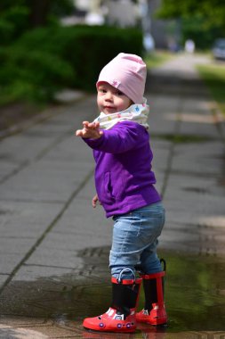Güneşli bir günde kaldırımdaki su birikintisinde oynayan kırmızı çizmeli ve renkli elbiseli tatlı bir kız. Çocukluk sevinci ve açık hava eğlencesi.