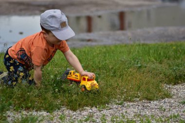 Şapkalı ve parlak elbiseli genç çocuk çimlerin üzerindeki su birikintisinin yanında sarı oyuncak kamyonla oynuyor. Çocuk odaklanmış ve meşgul. Doğada yaratıcılığı ve keşfi gösteren samimi açık hava anı.