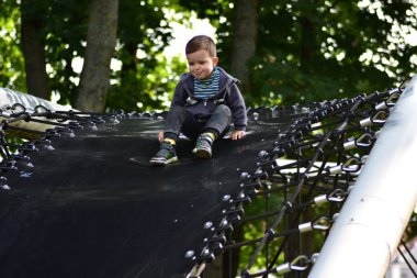 Kapüşonlu genç, modern oyun parkında trambolin benzeri dev bir halat yapısında oturuyor. Çocuk oyun sırasında dinlenirken hafifçe gülümser. Yeşil orman park ortamında açık hava eğlencesi.