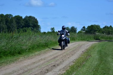 Motosikletçi, çakıllı kırsal yolda motosiklet turu yapan bir maceracı. Yeşil çimenli ve mavi gökyüzü olan kırsal alan. Özgürlük kavramı, yaz gezisi ve kırsal seyahat yaşam tarzı..