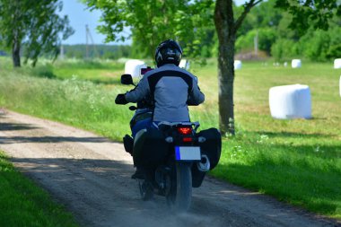 Motosikletçi, çakıllı kırsal yolda motosiklet turu yapan bir maceracı. Yeşil çimenli ve mavi gökyüzü olan kırsal alan. Özgürlük kavramı, yaz gezisi ve kırsal seyahat yaşam tarzı..