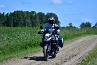 Motosikletçi, çakıllı kırsal yolda motosiklet turu yapan bir maceracı. Yeşil çimenli ve mavi gökyüzü olan kırsal alan. Özgürlük kavramı, yaz gezisi ve kırsal seyahat yaşam tarzı..