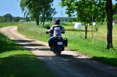Motosikletçi, çakıllı kırsal yolda motosiklet turu yapan bir maceracı. Yeşil çimenli ve mavi gökyüzü olan kırsal alan. Özgürlük kavramı, yaz gezisi ve kırsal seyahat yaşam tarzı..