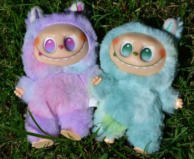 Kabarık yeşil ve pembe canavar kostümlü iki oyuncu Labubu oyuncak figürü ve muzip bir gülümseme..