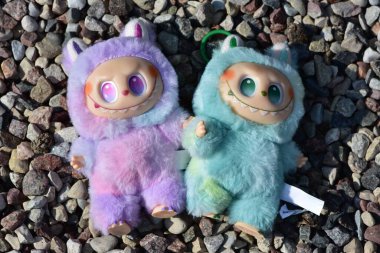 Kabarık yeşil ve pembe canavar kostümlü iki oyuncu Labubu oyuncak figürü ve muzip bir gülümseme..