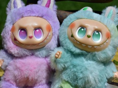Kabarık yeşil ve pembe canavar kostümlü iki oyuncu Labubu oyuncak figürü ve muzip bir gülümseme..