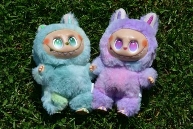 Kabarık yeşil ve pembe canavar kostümlü iki oyuncu Labubu oyuncak figürü ve muzip bir gülümseme..