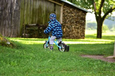 Kırsal kesimde arka bahçede bisiklete binen küçük bir çocuk. Yağmura dayanıklı kıyafetler giyen çocuklar güneşli bir günde dışarıda eğlenmekten ve fiziksel aktivitelerden hoşlanırlar. Yüzü yok.