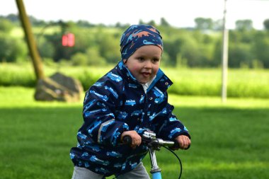 Ağzı açık genç bir çocuk kırsaldaki bir çimenlikte dengeli bisiklete biniyor. Yağmura dayanıklı kıyafetler giyen çocuklar güneşli bir günde dışarıda eğlenmekten ve fiziksel aktivitelerden hoşlanırlar..
