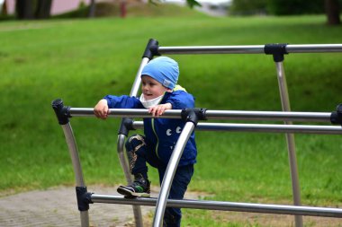 Mavi ceketli ve şapkalı genç çocuk modern metal oyun parkına tırmanıyor. Gülümseyen çocuk yeşil parkın dışında eğleniyor, heyecan ve özgürlüğü ifade ediyor..