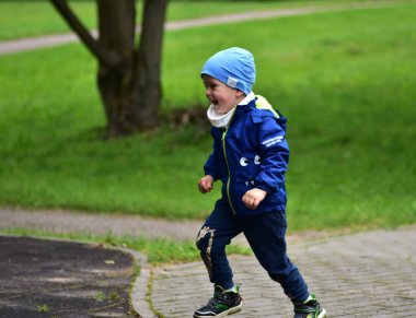 Yeşil parktaki asfalt yolda mavi ceketli ve şapkalı enerjik genç çocuk neşeyle koşuyor. Küçük çocuk dışarıda eğleniyor, doğada mutluluğu ve aktif yaşam tarzını ifade ediyor..