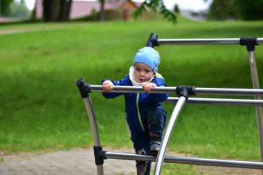 Mavi ceketli ve parktaki metal parmaklıklara tırmanan, kendinden emin bir şekilde ileriye bakan küçük bir çocuk. Yürüyen çocuk aktif olarak açık havada oyun oynuyor, motor becerilerini geliştiriyor ve parkta maceranın tadını çıkarıyor..