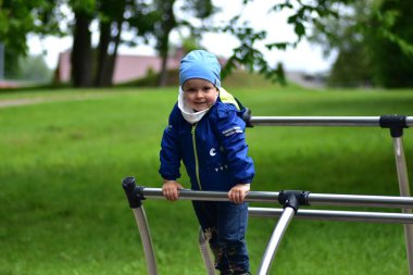 Gülümseyen genç çocuk metal oyun parkı ekipmanlarının üzerinde duruyor, mavi ceket ve şapka giyiyor. Parkta neşeli ve bağımsızlığını ifade eden kendinden emin çocuk dışarıda oynamaktan zevk alıyor..