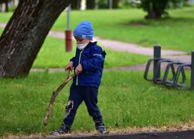 Mavi ceketli ve şapkalı genç yeşil parkta büyük sopalarla oynuyor. Çocuk yaratıcı bir açık hava oyunuyla meşgul, doğal materyalleri keşfediyor ve yaratıcılığını ve merakını ifade ediyor..