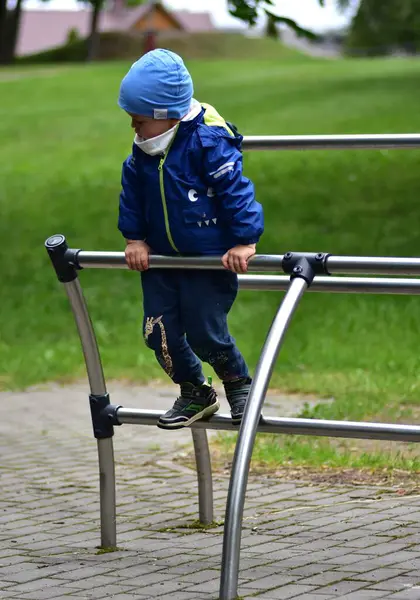 Metal oyun parkında dengede duran, dikkatlice bakan genç bir çocuk. Mavi ceketli ve şapkalı çocuk yemyeşil park ortamında asfalt bir alanda meydan okuyarak eğleniyor..