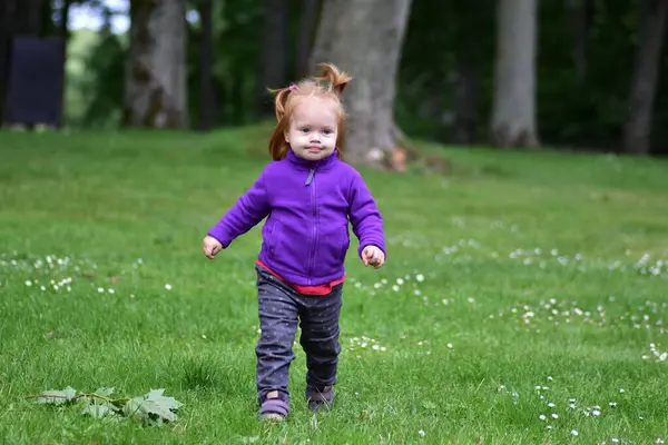 Mor ceketli kızıl saçlı küçük kız yeşil çimlerin üzerinde tek başına geziyor, açık havayı keşfediyor. Neşeli, bağımsız çocukluk anı doğal ışıkla yakalanır, özgürlüğü ve merakı sembolize eder.