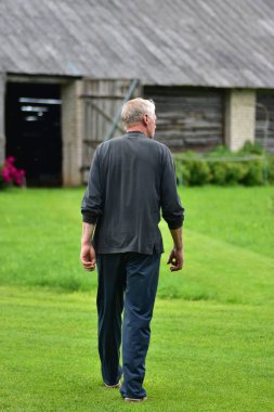 Yeşil çimlerde tek başına yürüyen, sıradan giysili yaşlı bir adam, arkadan görülüyor. Huzurlu kırsal atmosfer, yalnızlık, tefekkür, yaşlanma ve basit kırsal yaşam kavramı..