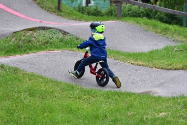 Yeşil parkta asfalt yolda kırmızı dengeli bisiklete binen bir çocuğun arka görüntüsü. Çocuk bisikletçilik becerilerini bağımsız olarak keşfediyor. Çocukluk macerası, büyüme ve açık hava aktivitesi kavramı.
