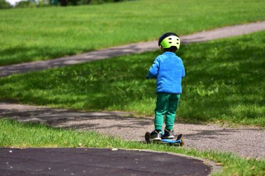 Mavi ceketli küçük bir çocuk yeşil kask takmış, tek başına park yolunda scooter sürüyor. Bağımsızlığı ve eğlenceyi teşvik eden yaz etkinliği.