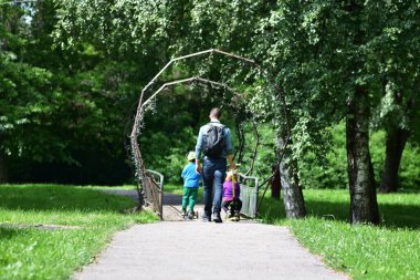 Babam sırt çantasıyla ve iki çocuğuyla yemyeşil parktaki köprüde scooter sürüyor. Hafta sonu aile aktiviteleri açık havada. Babam yazın çocuklarla parkta vakit geçiriyor..