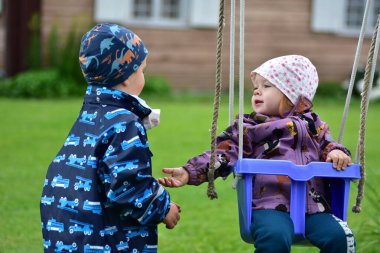 İki küçük çocuk salıncak seti üzerinde dışarıda etkileşime geçiyorlar. Kız oturuyor, oğlan ayakta, renkli ceketler ve şapkalar giyiyor. Kırsalda arkadaşlık, oyun zamanı ve çocukluk sevinci kavramı.