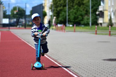 Güneşli bir günde mavi scooter kullanan bir çocuk konut alanında asfalt yolda. Çizgili kapüşonlu ve şapkalı genç aktif açık hava oyunlarının keyfini çıkarıyor, çevreyi keşfediyor ve neşe içinde hareket becerilerini geliştiriyor.