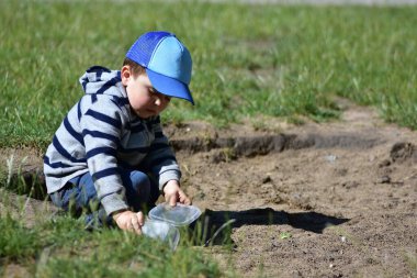 Genç çocuk güneşli bir günde kum havuzunda plastik konteynırla oynuyor. Çizgili kapüşonlu ve mavi şapkalı çocuk hayal ürünü açık hava oyunlarından zevk alıyor, merakı, öğrenmeyi ve çocukluk eğlencesini sembolize ediyor.