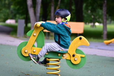 Çocuk oyun parkında motosiklet şeklinde yeşil kapüşonlu ve kask takan bir yaylı rockçının üzerinde zıplıyor. Neşeli sahne, hareketler, heyecan ve çocukluk oyunculuğunu renkli açık havada yakalar..