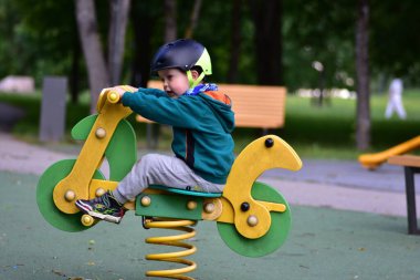 Çocuk oyun parkında motosiklet şeklinde yeşil kapüşonlu ve kask takan bir yaylı rockçının üzerinde zıplıyor. Neşeli sahne, hareketler, heyecan ve çocukluk oyunculuğunu renkli açık havada yakalar..