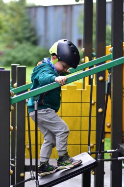 Genç çocuk modern oyun parkı köprüsüne tırmanıyor, güvenlik kaskı takıyor ve yeşil kapüşonlu giyiyor. Açık hava oyununda dengeyi, odaklanmayı ve kendine güveni yakalayan, sağlıklı çocukluk gelişimini teşvik eden aksiyon çekimi.