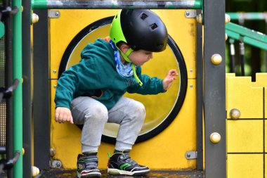 Çocuk oyun parkındaki dairesel tünelde çömelmiş, güvenlik kaskı ve yeşil kapüşonlu kazak giyiyor. Meraklı ve oyuncu ifade, keşfin sembolü, eğlence ve güvenli ortamda yaratıcı oyun.