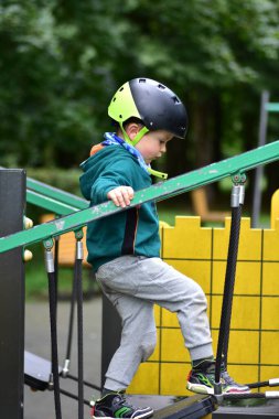 Genç çocuk modern oyun parkı köprüsüne tırmanıyor, güvenlik kaskı takıyor ve yeşil kapüşonlu giyiyor. Açık hava oyununda dengeyi, odaklanmayı ve kendine güveni yakalayan, sağlıklı çocukluk gelişimini teşvik eden aksiyon çekimi.