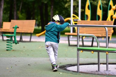 Genç bir çocuk oyun parkında tek başına, kasklı ve yeşil kapüşonlu bir şey giyiyor. Sahne, renkli park ortamındaki aktif açık hava oyunlarında bağımsızlık, hareket ve merakı vurguluyor.